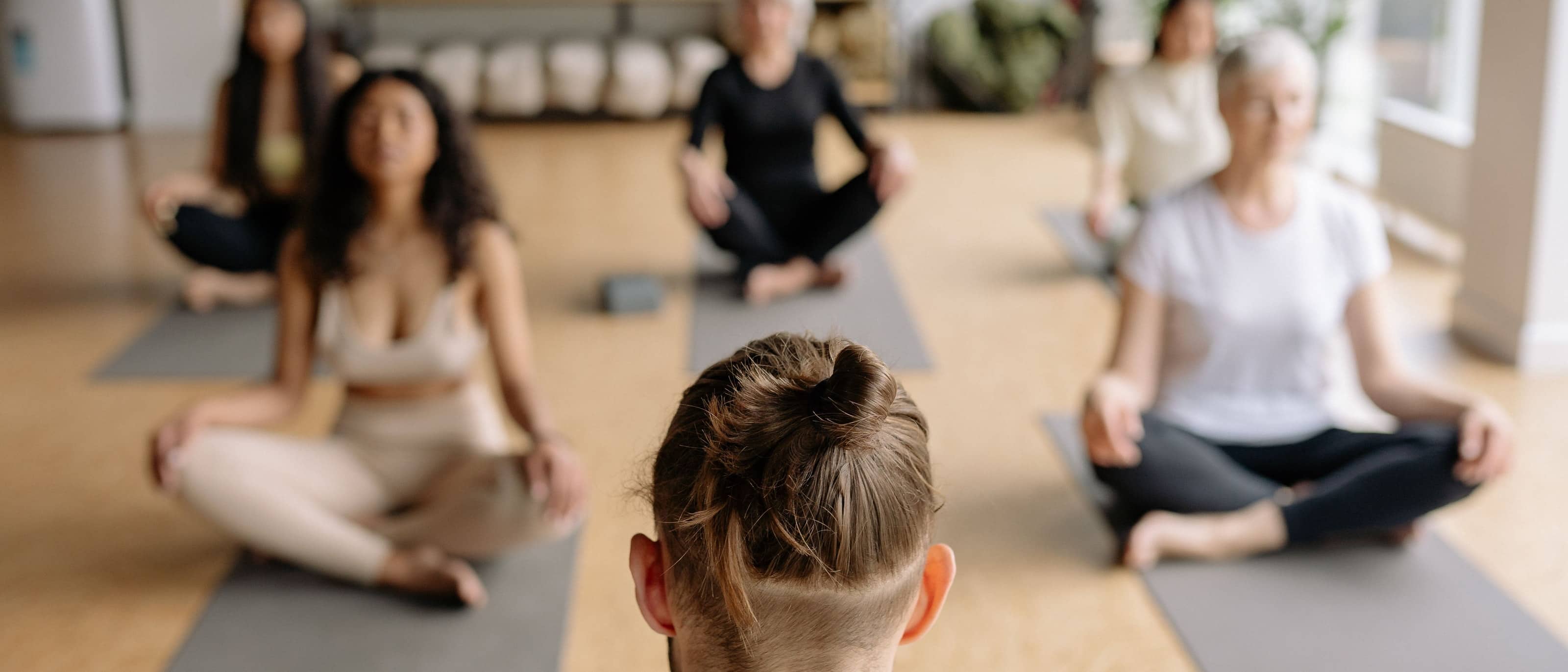A yoga class seen from behind the instructor: five people sitting cross-legged on mats in a bright studio with a cork floor, eyes closed.
