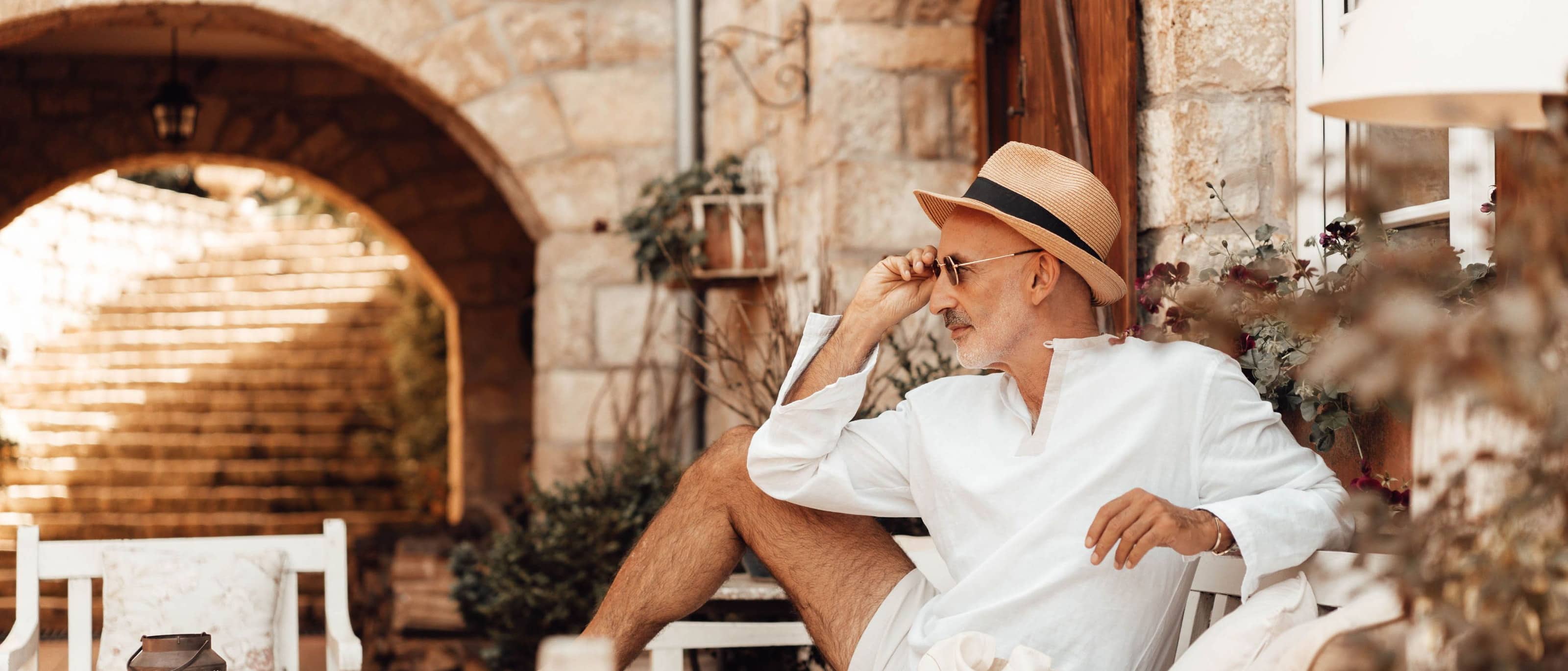 A man in his sixties in a white linen shirt and straw hat, seated on a bench in a shaded stone courtyard, looking out past the camera into warm afternoon light.
