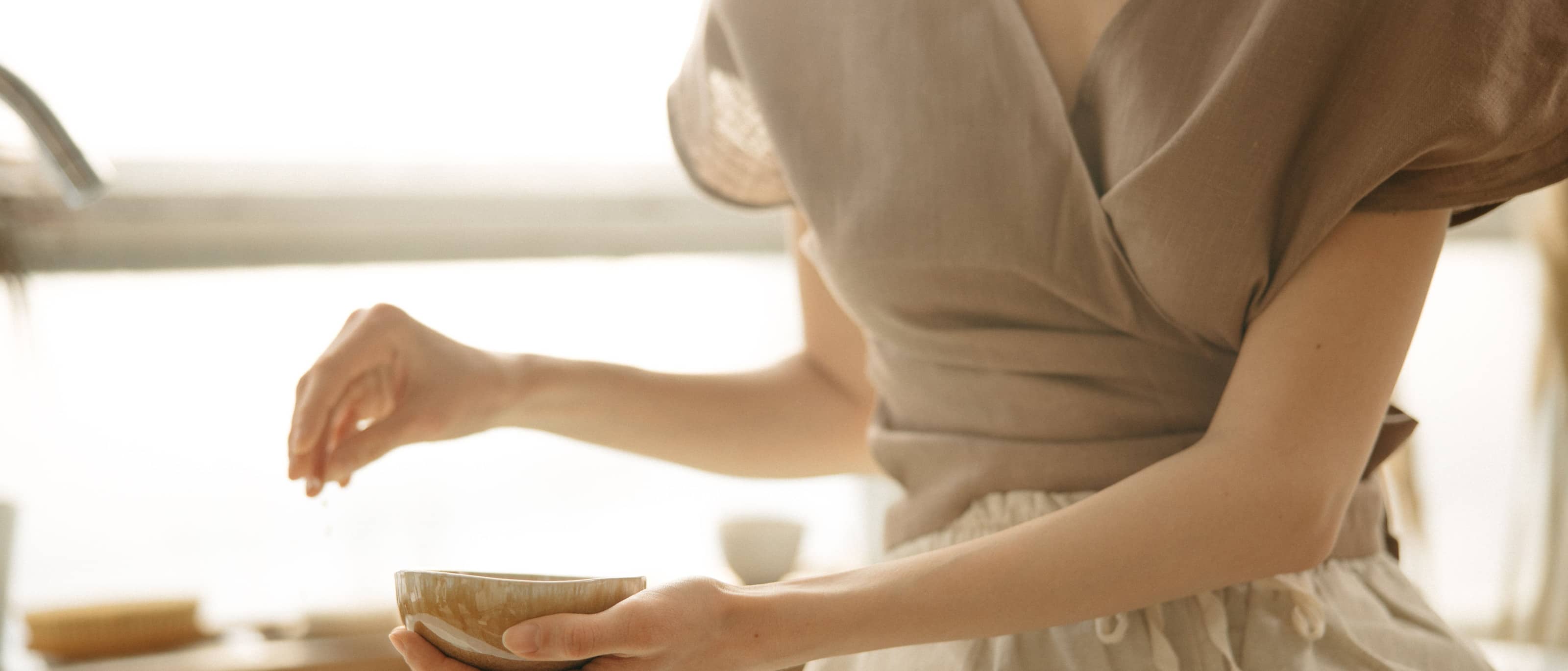 A woman in soft linen holding a small ceramic bowl, seen at a table in diffused daylight.