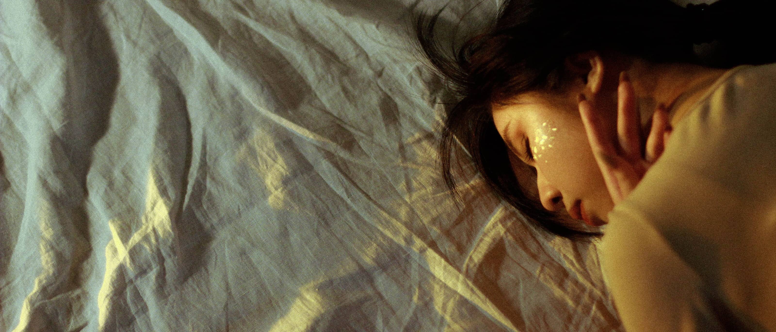 A woman asleep on her side, dappled morning light falling across crumpled cream linen.