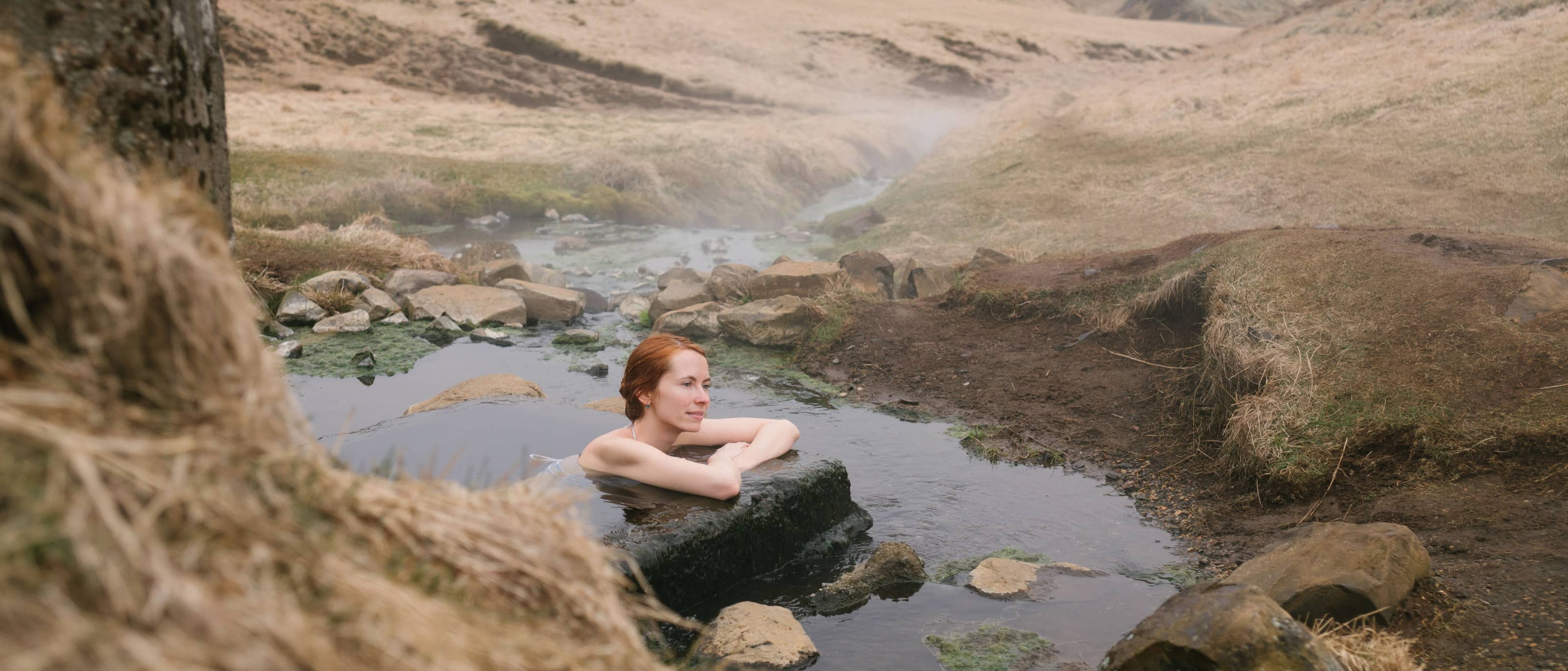 A woman sitting shoulder-deep in a natural thermal pool, steam drifting off the water, dry ochre hills rising on both sides.
