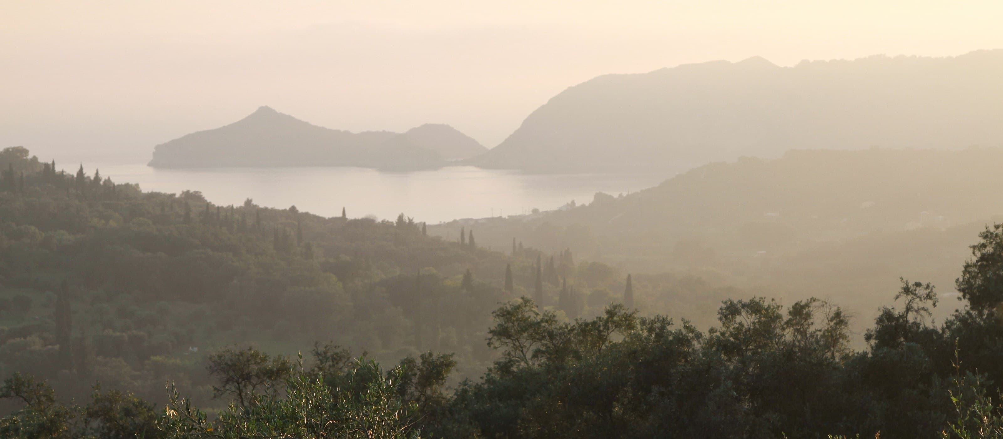 Layered headlands receding into warm haze above a still bay, cypress and scrub standing on the near slope in low sun.