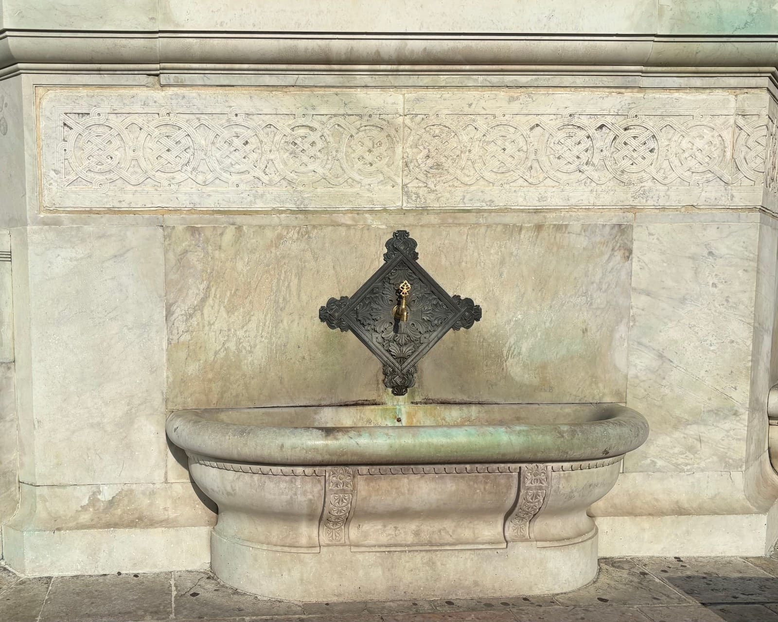 A marble wall fountain — a carved interlace frieze above a panelled slab, a bronze spout, and a thread of water falling into a heavy carved basin.
