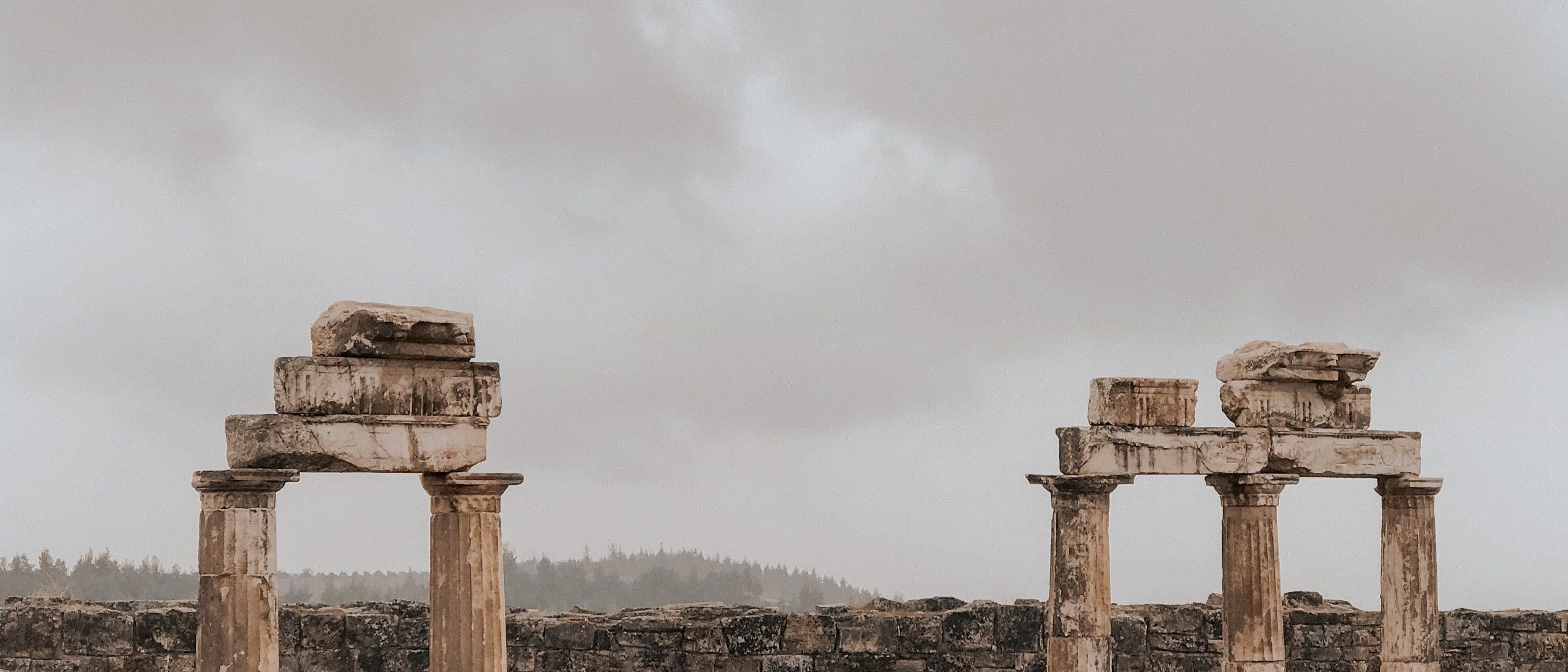 Two groups of ruined limestone columns standing against a pale overcast sky at Hierapolis, a low masonry wall and a hazy pine ridge behind them.