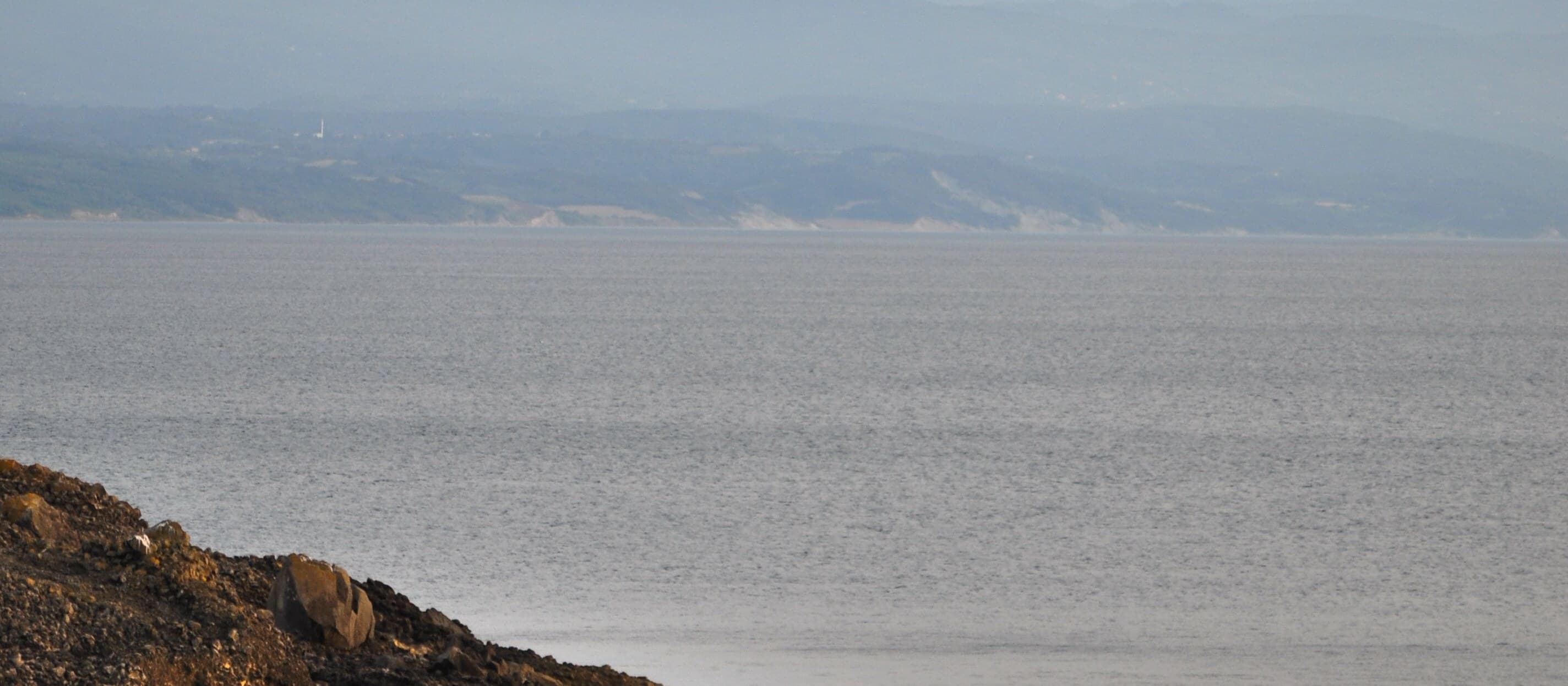 A rocky headland entering calm water on the left, a far shore lying low under a pale hazy sky.