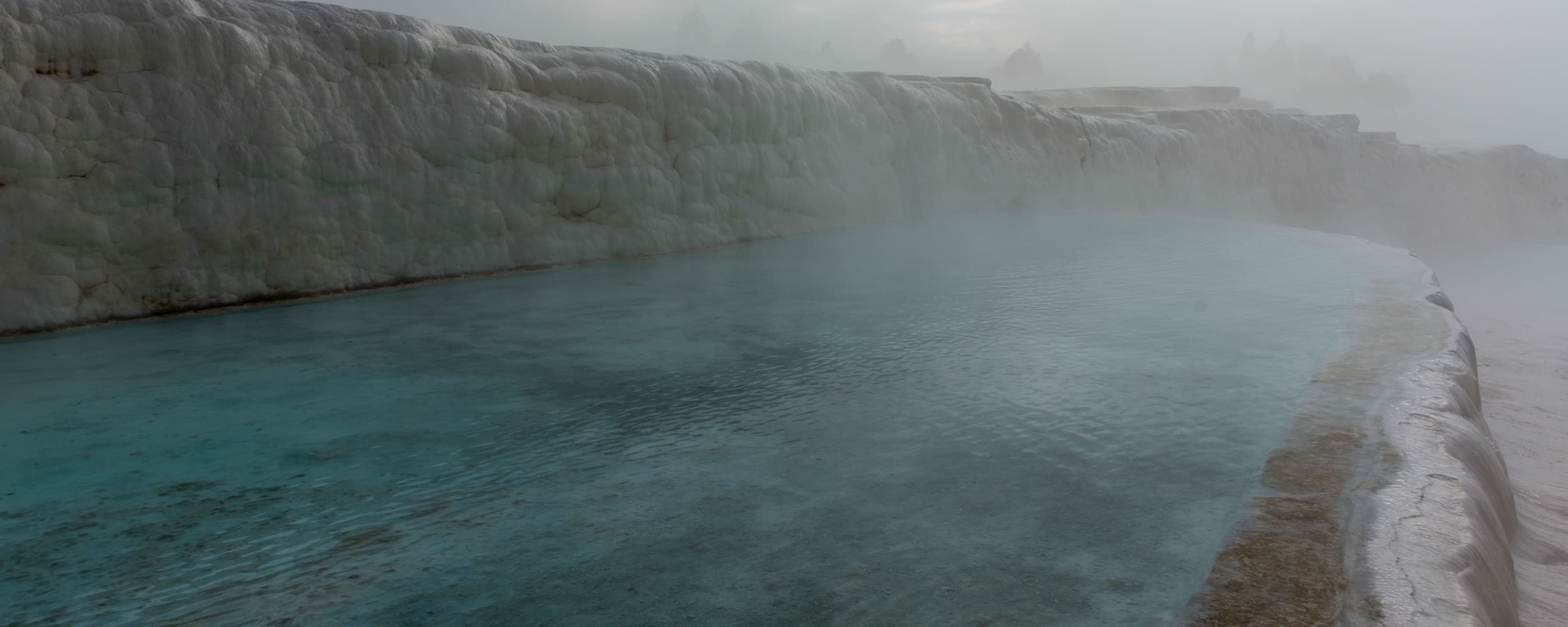 Steam drifting over the turquoise thermal water held behind a white travertine wall at Pamukkale.