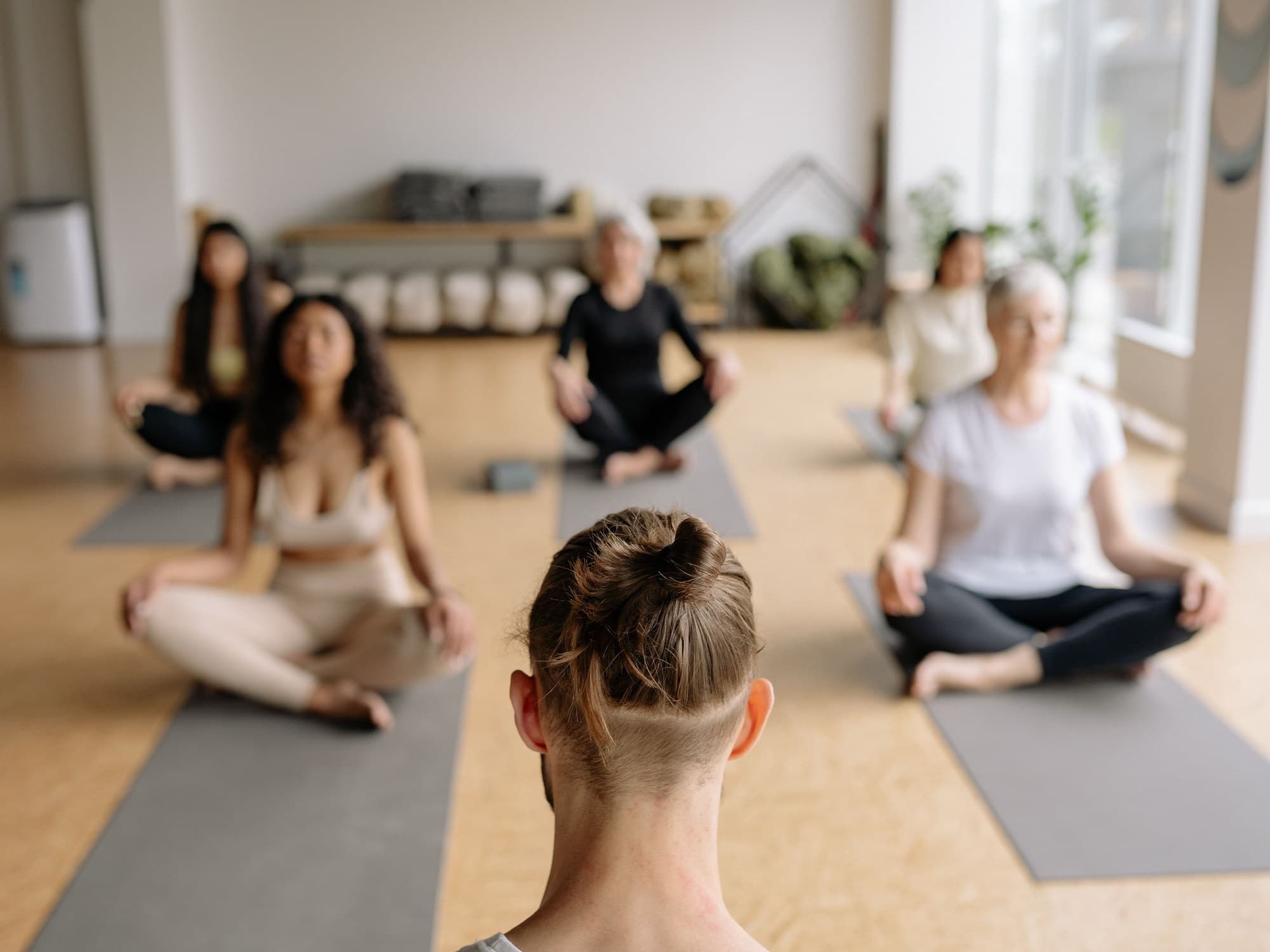 A yoga class seen from behind the instructor: five people sitting cross-legged on mats in a bright studio with a cork floor, eyes closed.