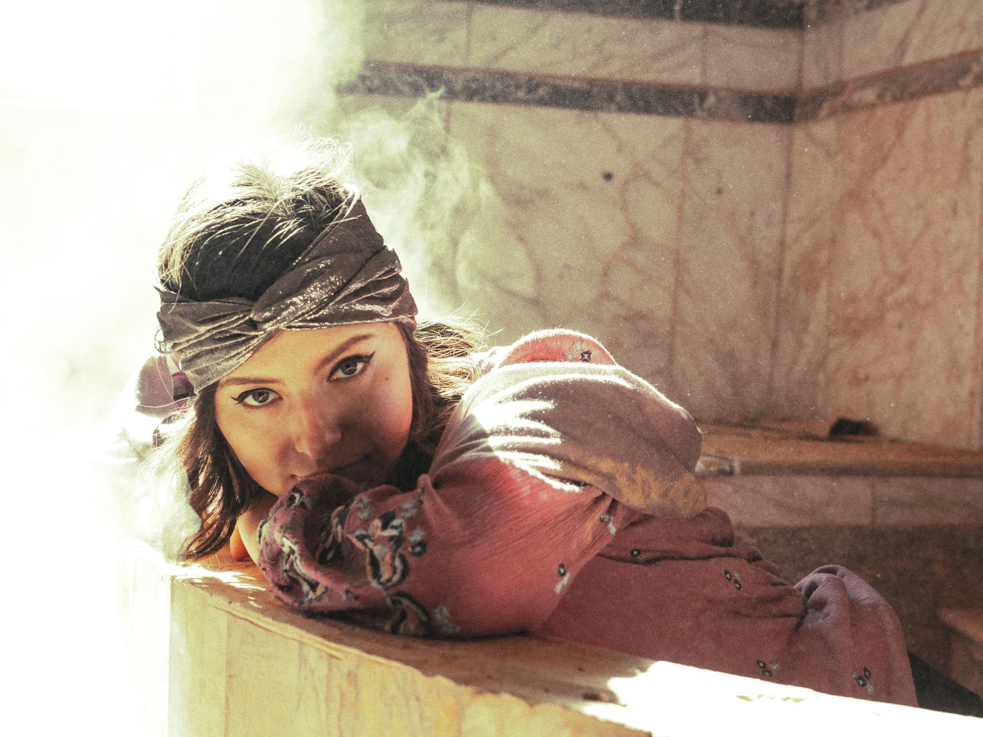 A woman resting against warm marble in a hammam, wrapped in a rose-coloured pestemal, steam and daylight behind her.