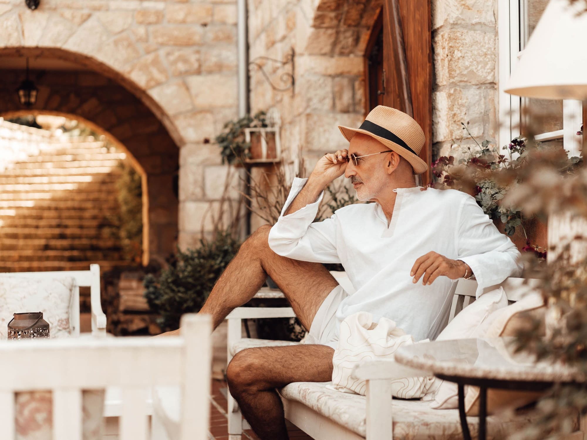 A man in his sixties in a white linen shirt and straw hat, seated on a bench in a shaded stone courtyard, looking out past the camera into warm afternoon light.