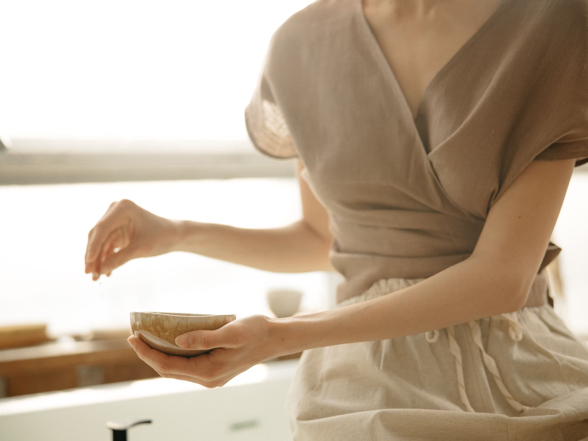 A woman in soft linen holding a small ceramic bowl, seen at a table in diffused daylight.
