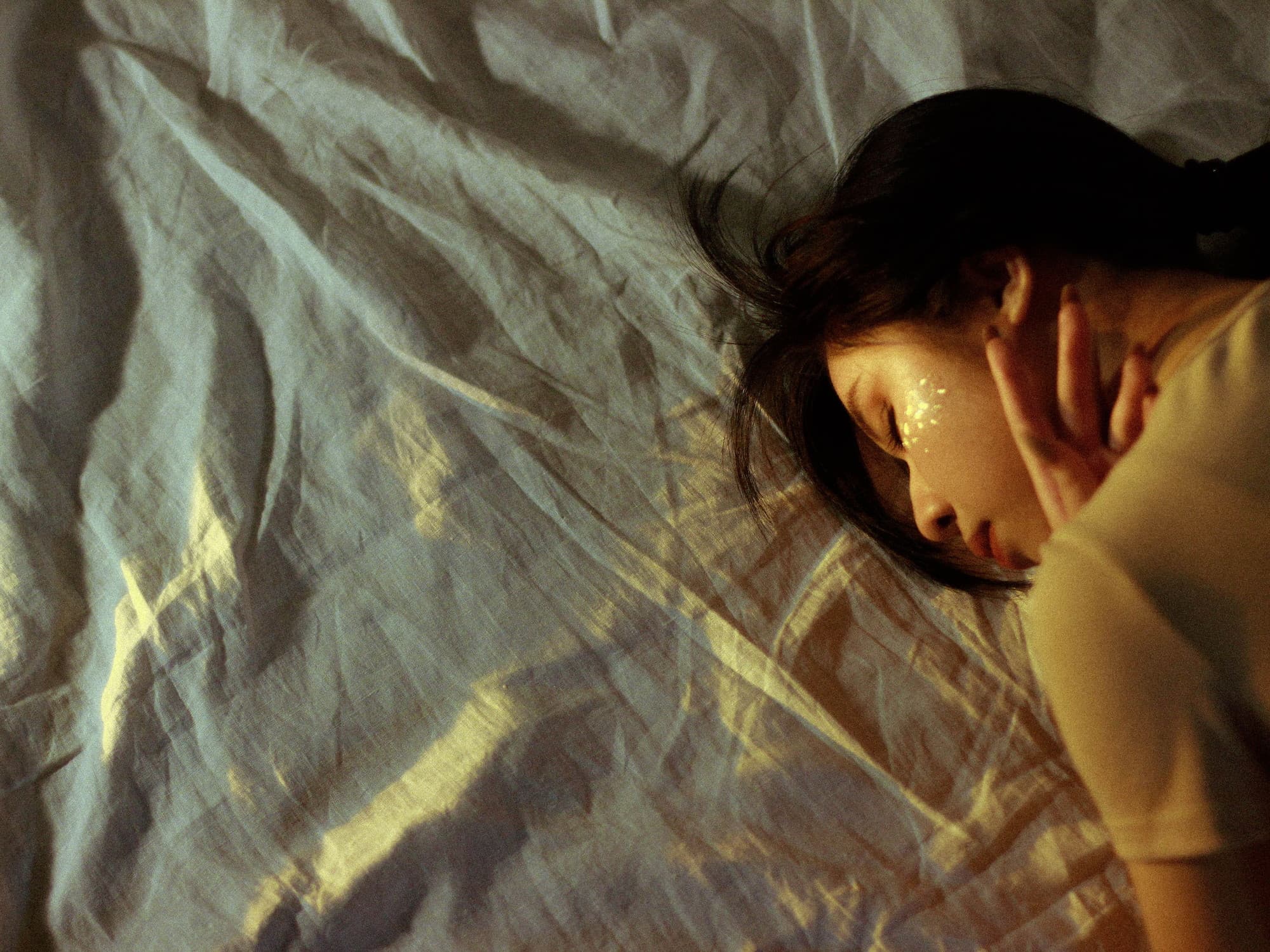 A woman asleep on her side, dappled morning light falling across crumpled cream linen.
