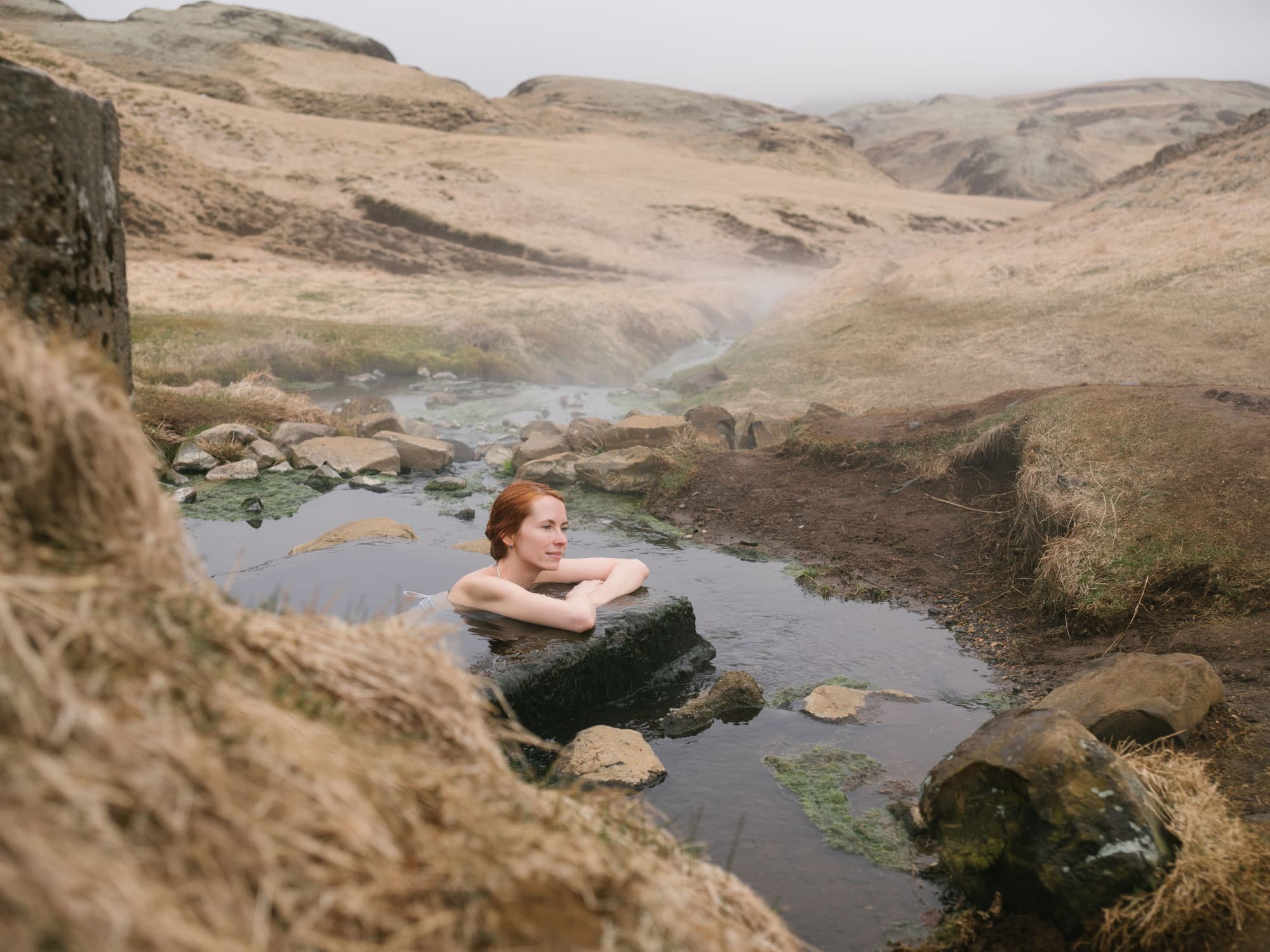 A woman sitting shoulder-deep in a natural thermal pool, steam drifting off the water, dry ochre hills rising on both sides.