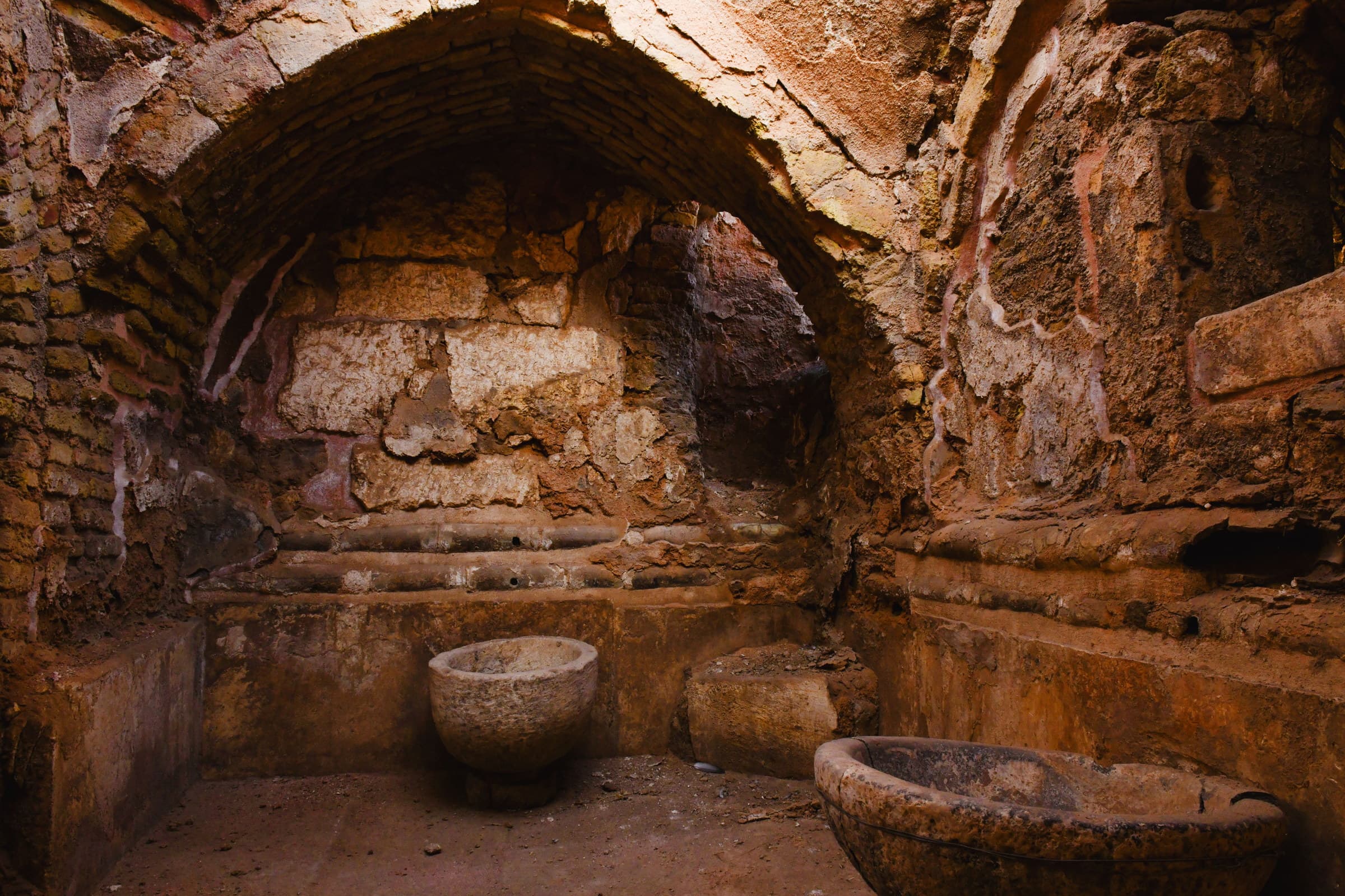 A vaulted stone bathing room at Harran — worn brick and plaster, a shallow ledge along the wall, and two carved basins on the floor in warm raking light.