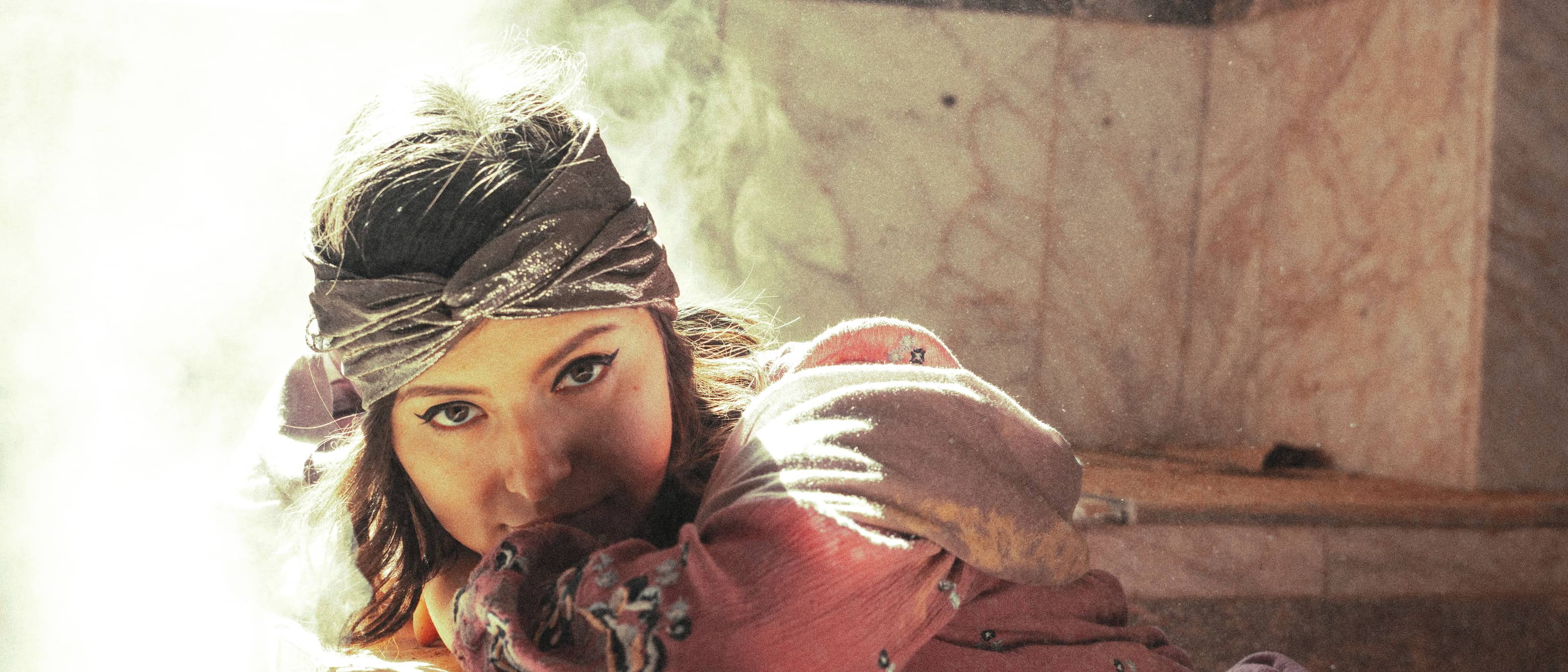 A woman resting against warm marble in a hammam, wrapped in a rose-coloured pestemal, steam and daylight behind her.