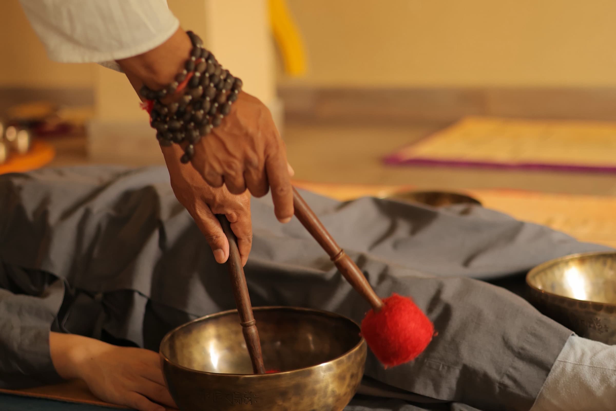 A practitioner's hand, wrist wrapped in mala beads, striking a brass singing bowl resting on a person lying covered on a mat.