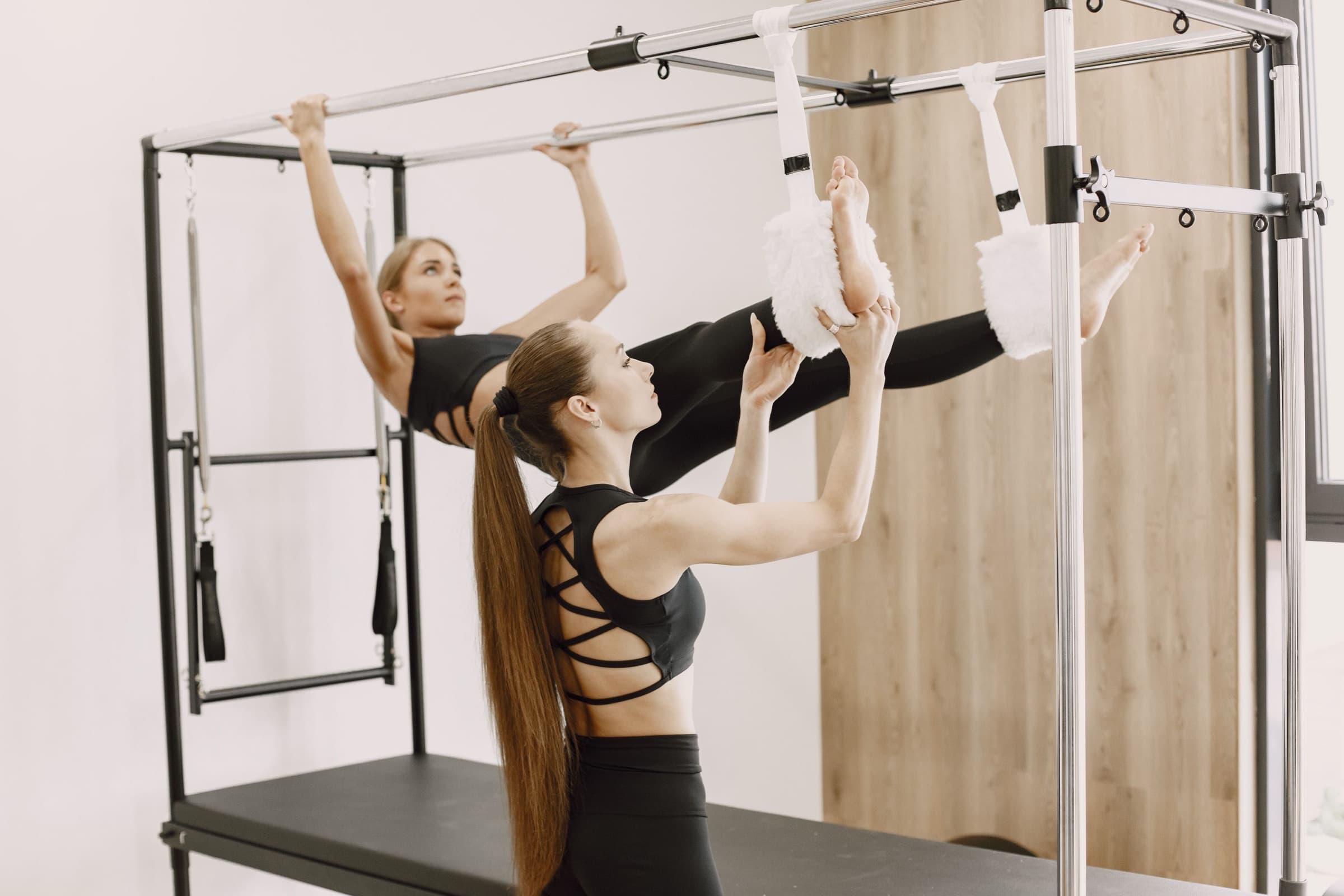 Two people working on pilates reformers in a pale studio with a timber wall.
