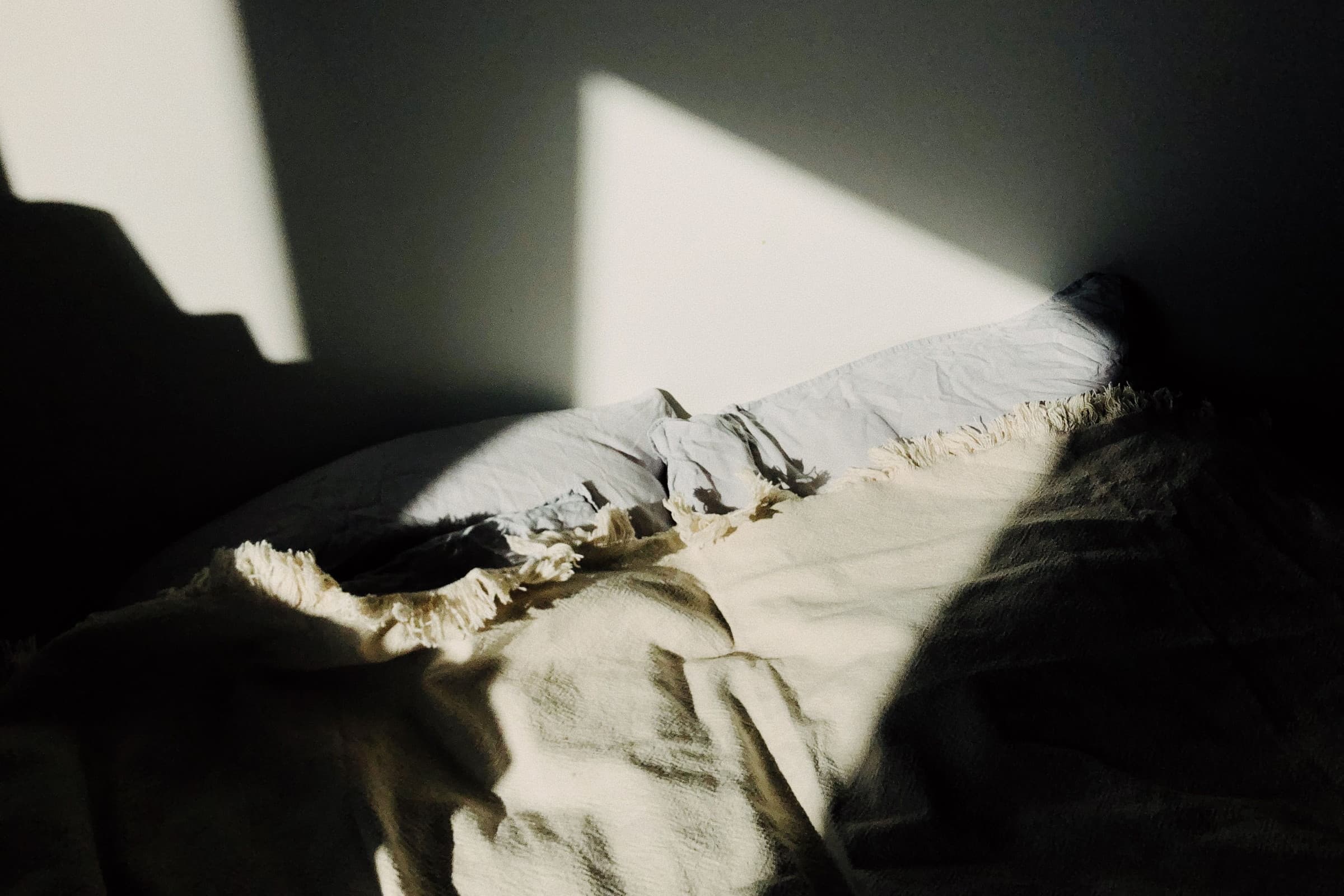Morning light broken into soft shapes across rumpled bed linen, the room beyond it still in shadow.