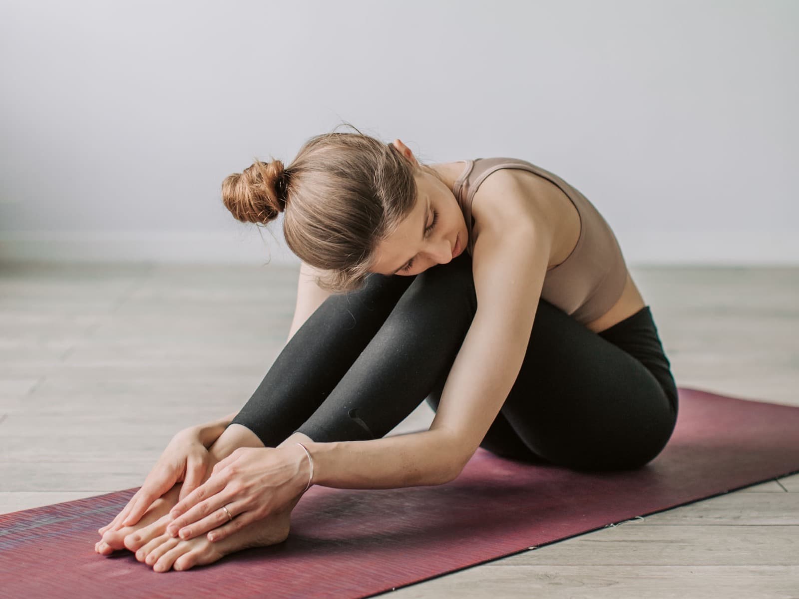 A person seated on a deep red mat in a bare room, folded forward over straight legs with hands at the feet.