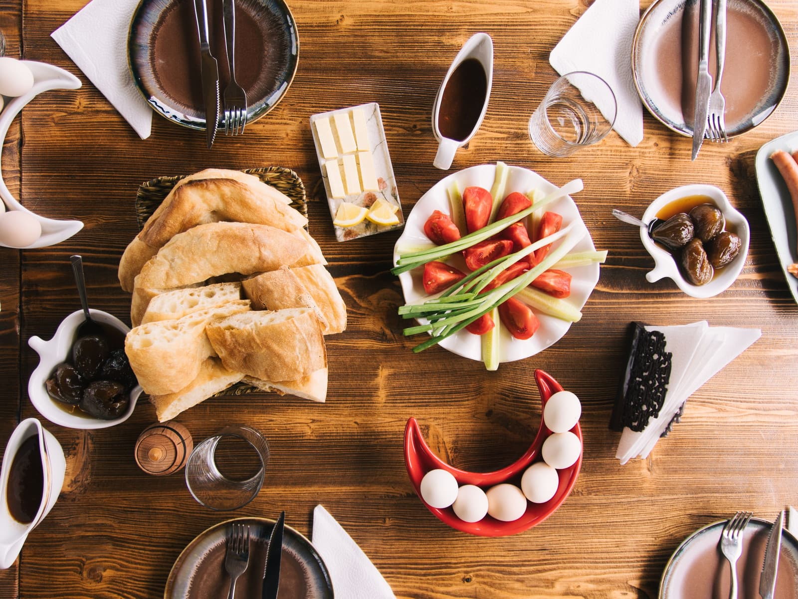 A breakfast table seen from above — bread, cheeses, olives, tomatoes and cucumber laid out across dark timber.