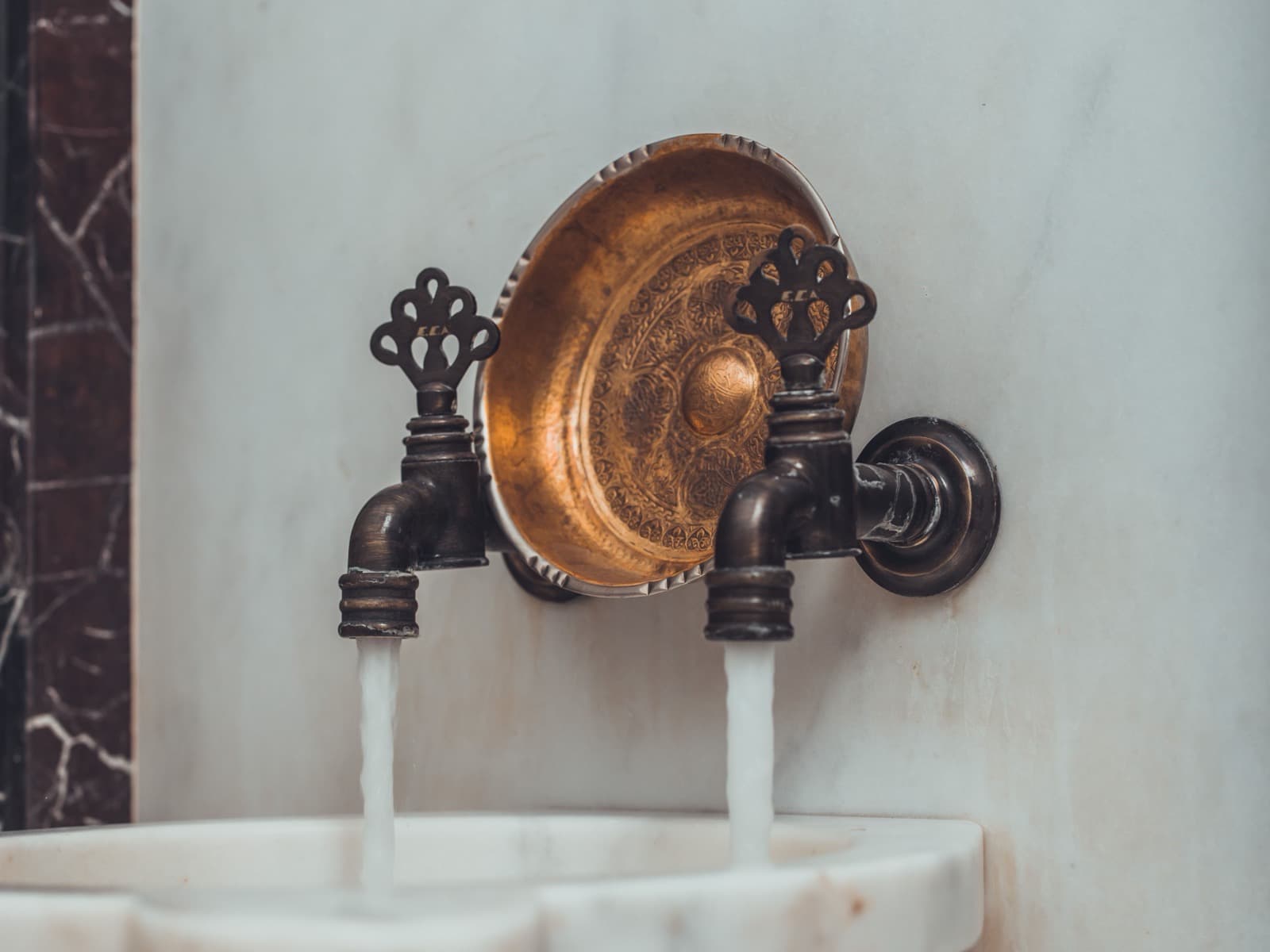Two ornate brass taps running into a scalloped white marble basin in a hammam, dark marble inlay at the edge.