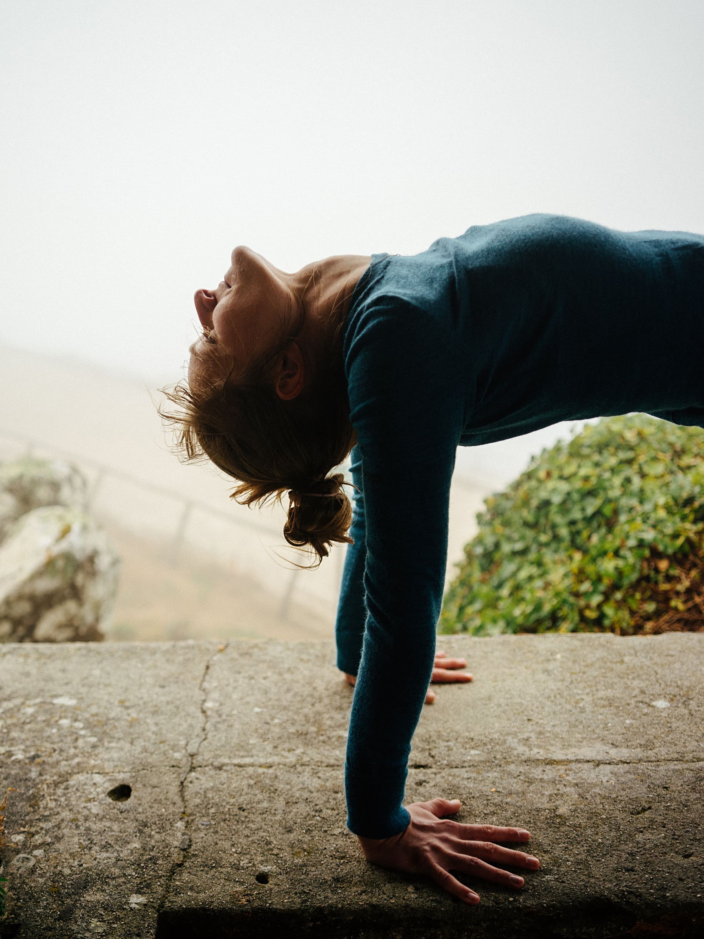 A woman in a downward-facing dog on a bare stone terrace, sea fog closing in behind her.